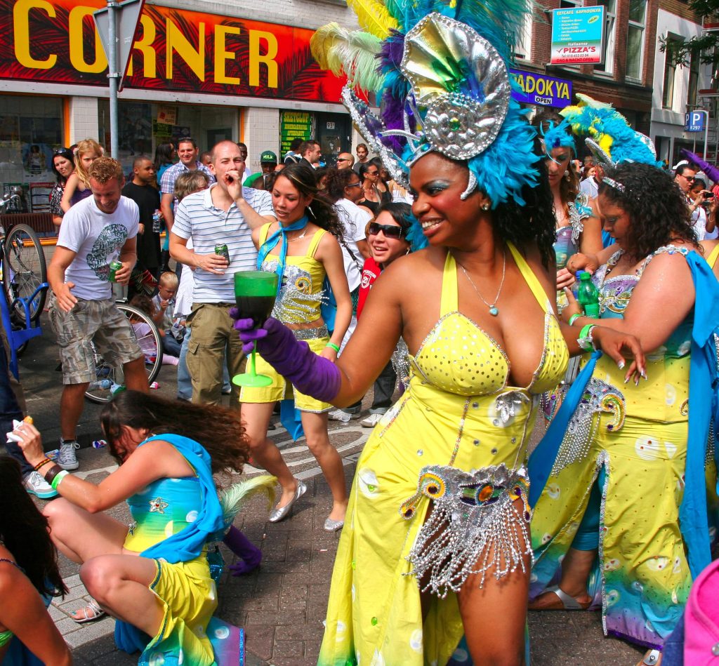 Woman in Carnival costume dancing down the streets at Rotterdam Zomercarnaval with a happy crowd in the background dancing along on the sidewalks.