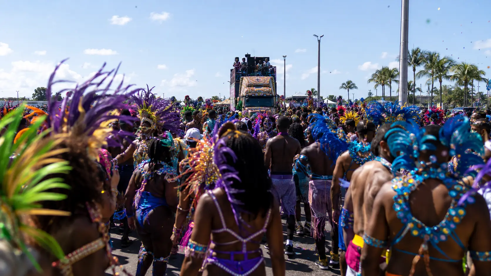 Miami Carnival Parade of Bands at the Miami-Dade County Fairgrounds with costumed masqueraders in colorful feathered costumes walking along an open parade route under a sunny blue sky.