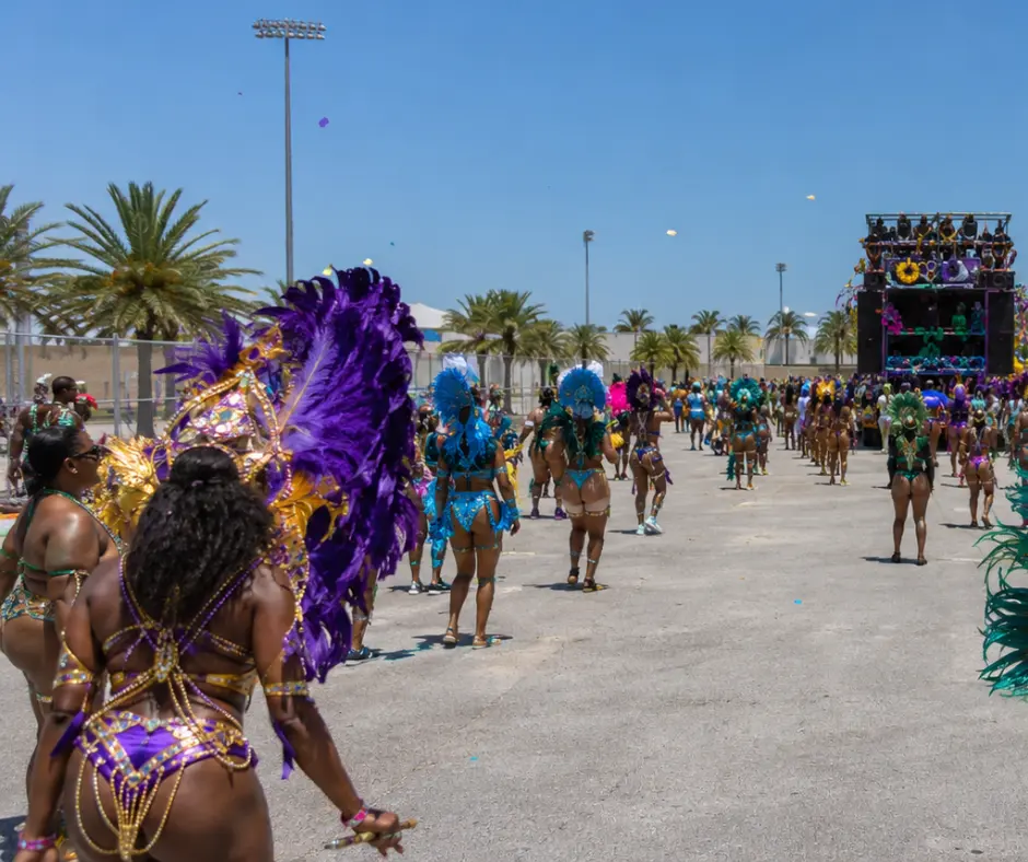 Masqueraders in purple, gold, and teal feathered Caribbean mas costumes dancing toward a decorated music truck at the Miami Carnival Parade of Bands, with palm trees and light poles visible under a clear blue sky
