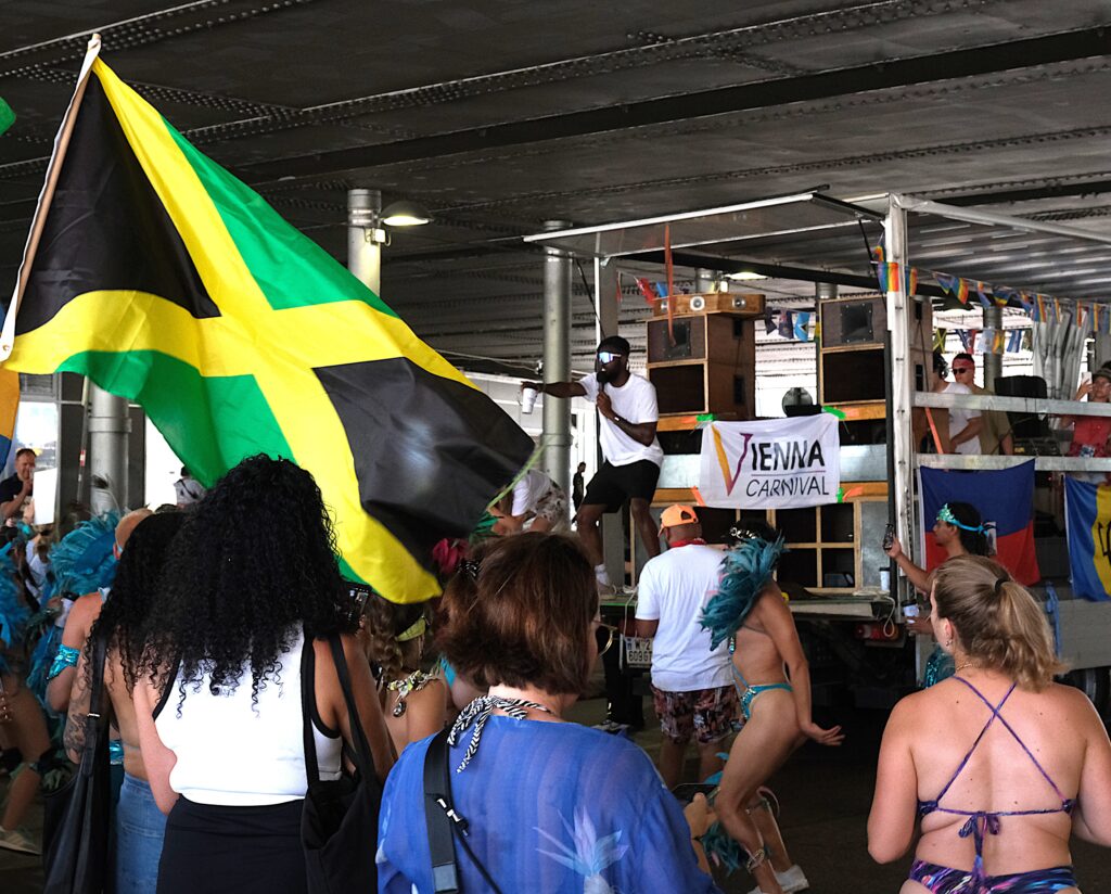 Man with microphone pointing to the crowd and hyping up revelers on the back of a soca truck at the Vienna Carnival Road March 2024, with a Jamaica flag visible.