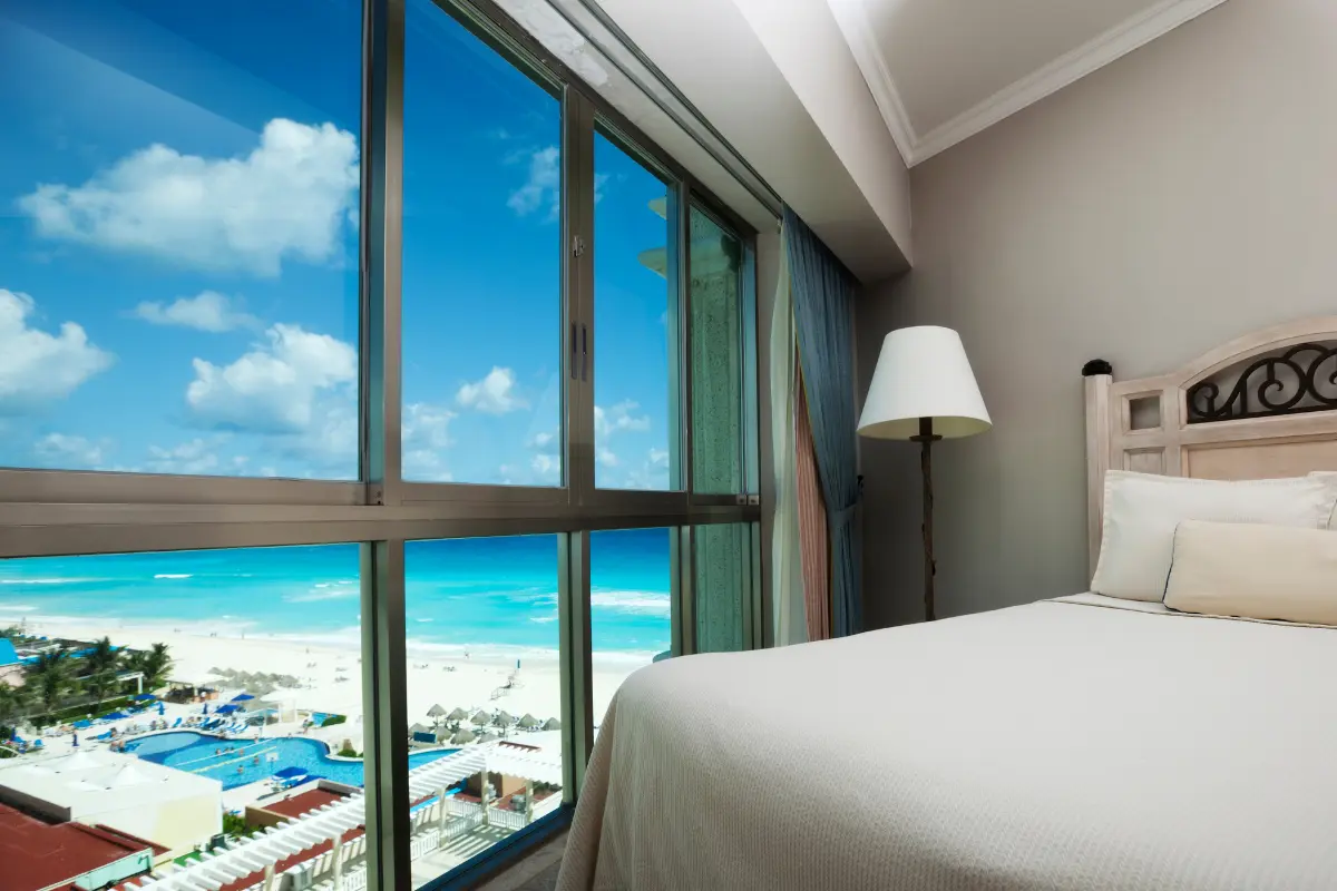 Bright Caribbean hotel room with a large ocean-view window overlooking a white sand beach and turquoise sea.