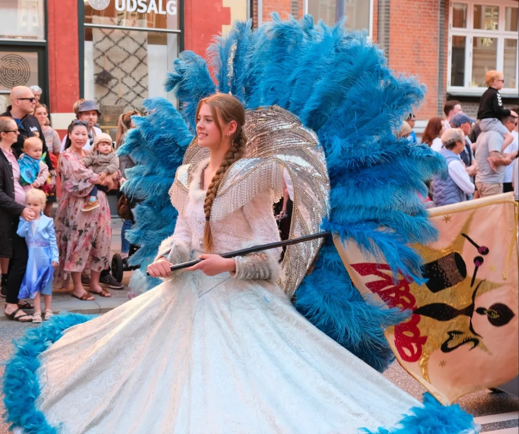 Performer in blue feather carnival costume during the Aalborg Carnival parade in Aalborg, Denmark with spectators watching from the street.