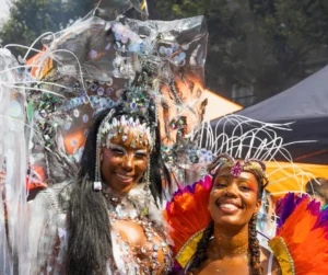 Two women smiling in elaborate carnival costumes with glitter makeup and headpieces, demonstrating the full experience of styling when learning how to get a carnival costume