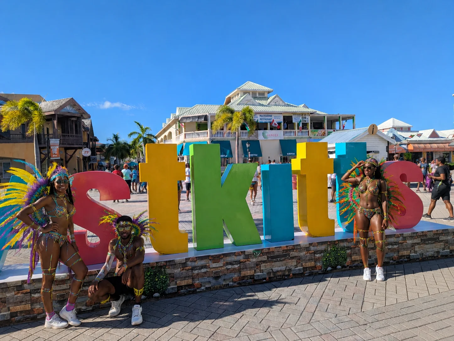 Real photo of the St. Kitts sign in Basseterre with AI-generated Sugar Mas masqueraders posing for photos before joining the road during one of the featured events on the list of Caribbean carnivals.
