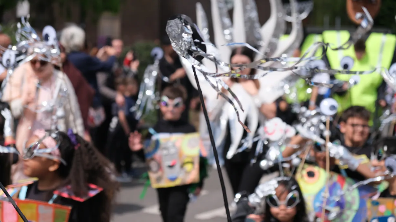 Participants in silver metallic and black eye-themed costumes carrying handmade props and placards through a street carnival parade in December