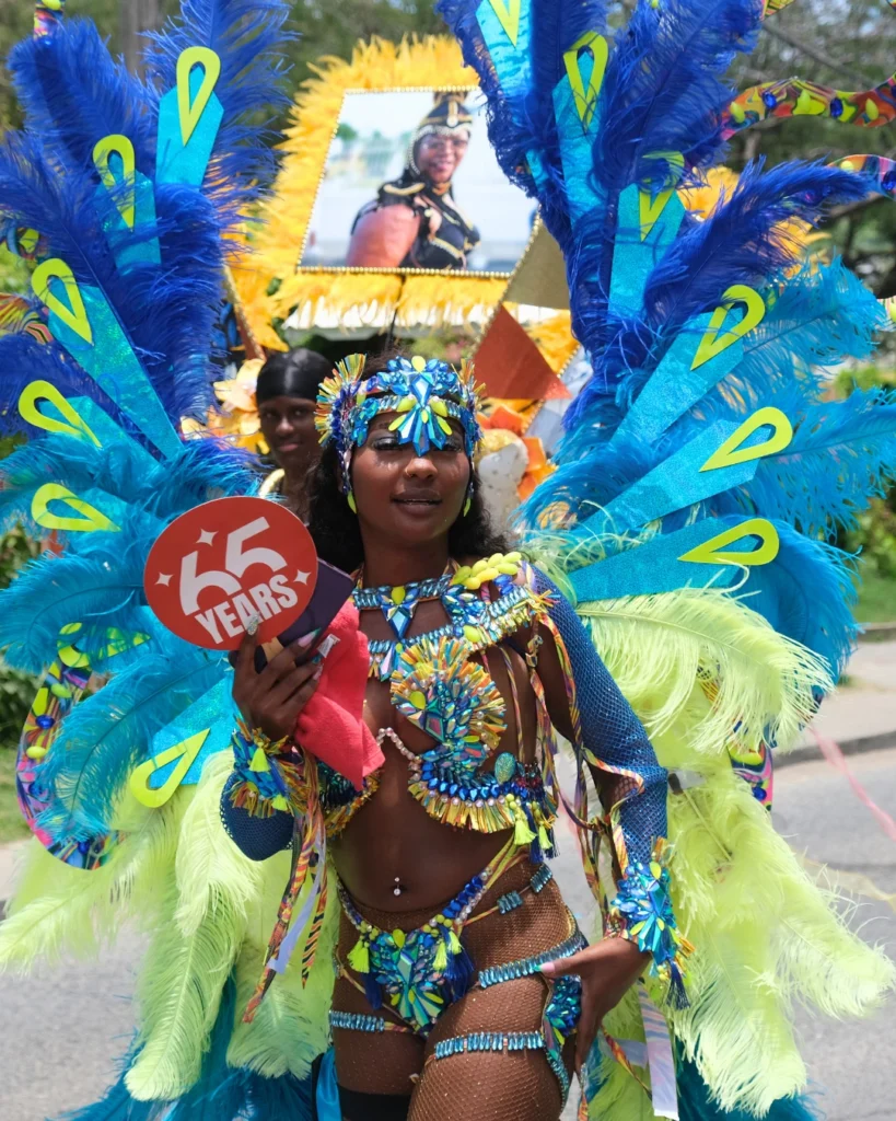 Masquerader in blue and lime carnival costume holding anniversary sign, featured as a carnival travel resource