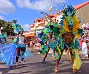 Dancers from different dance troupes on parade for the perform in Rue du Président Kennedy, Marigot during the Marigot Carnival or "Carnaval de St. Martin", an annual event / celebration in Saint-Martin. The dancers wear brightly colored costume decorated with flamboyant feathers on the headdress. The dancer on the left is holding out "wings of isis" which have become a popular modern addition to the traditional costumes.