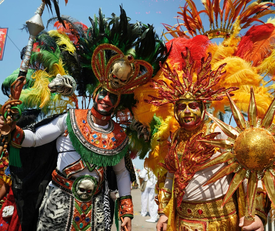 Two performers in elaborate South American carnival character costumes with feathered headdresses, skull masks, and full face paint at Barranquilla Carnival