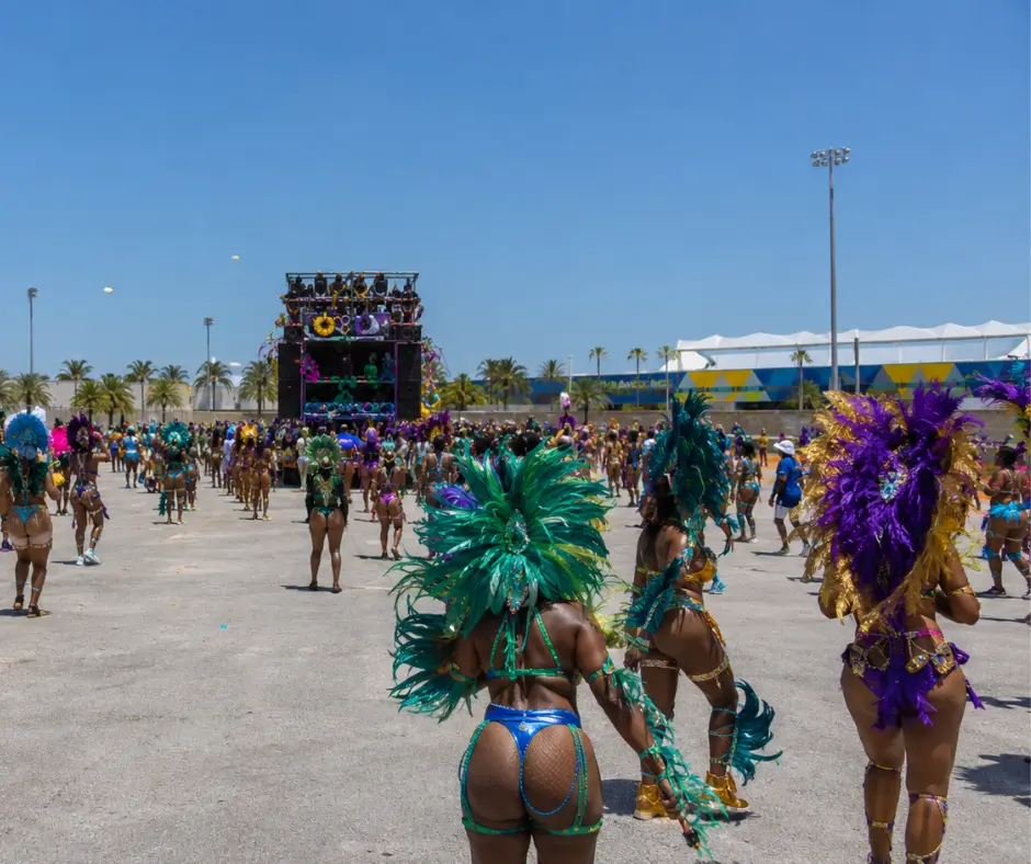 Thousands of masqueraders in green, purple, and gold feathered carnival costumes filling the Miami-Dade Fairgrounds during the Miami Carnival Parade of Bands, with confetti in the air and a music truck visible in the distance