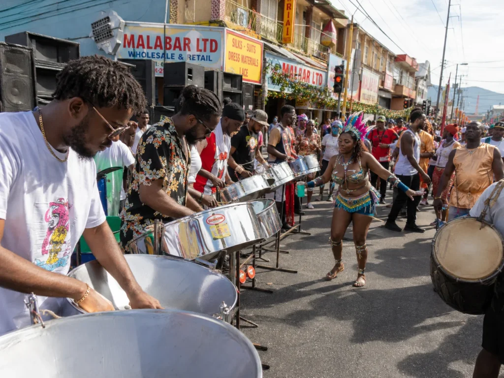 Steelpan musicians perform on a Trinidad street during Carnival as dancers, tassa drums, speaker boxes, and colorful storefronts celebrate the roots of soca music.