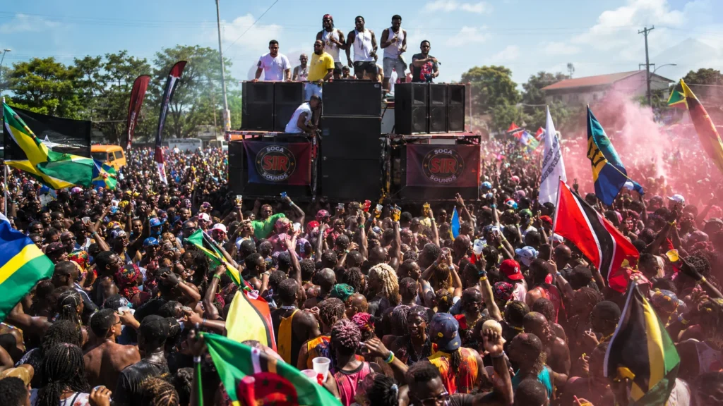 Massive Carnival crowd jumps behind a towering speaker truck with flags, powder, DJs, and performers celebrating the explosive energy of power soca music.