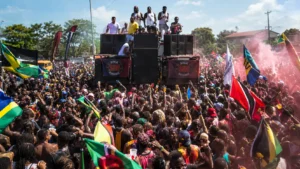 Massive Carnival crowd jumps behind a towering speaker truck with flags, powder, DJs, and performers celebrating the explosive energy of power soca music.