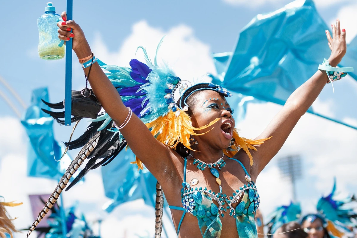 Masquerader in a turquoise feathered costume with gems raising her arms and waving a flag as she celebrates on the road at Caribbean Carnival.