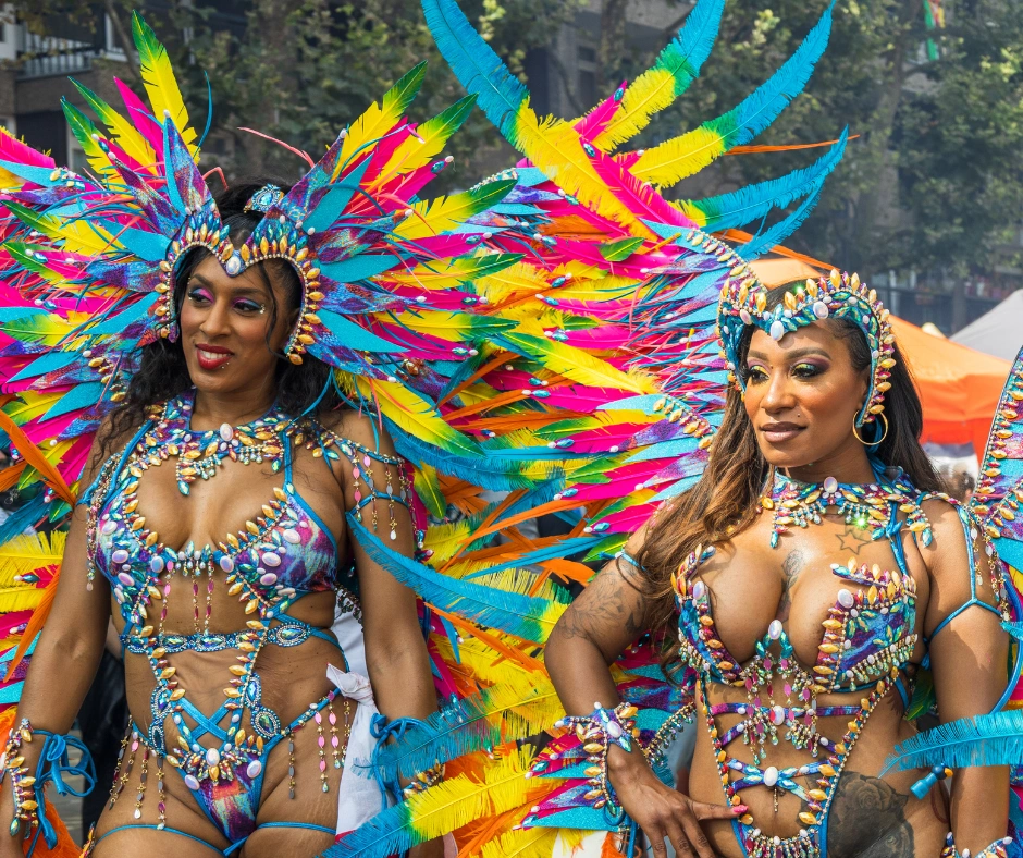Two masqueraders in rainbow feathered headdresses and shell-trimmed costumes on a tree-lined city street
