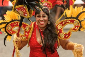 Smiling masquerader in a red sequinned costume with a gold and red jewelled headpiece, black and yellow feathers, and matching gem-covered hand pieces at Aruba Carnival