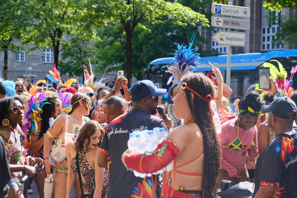 Crowd of masqueraders in bright feathered headpieces and gem-covered costumes filling a sunlit street during Munich Carnival