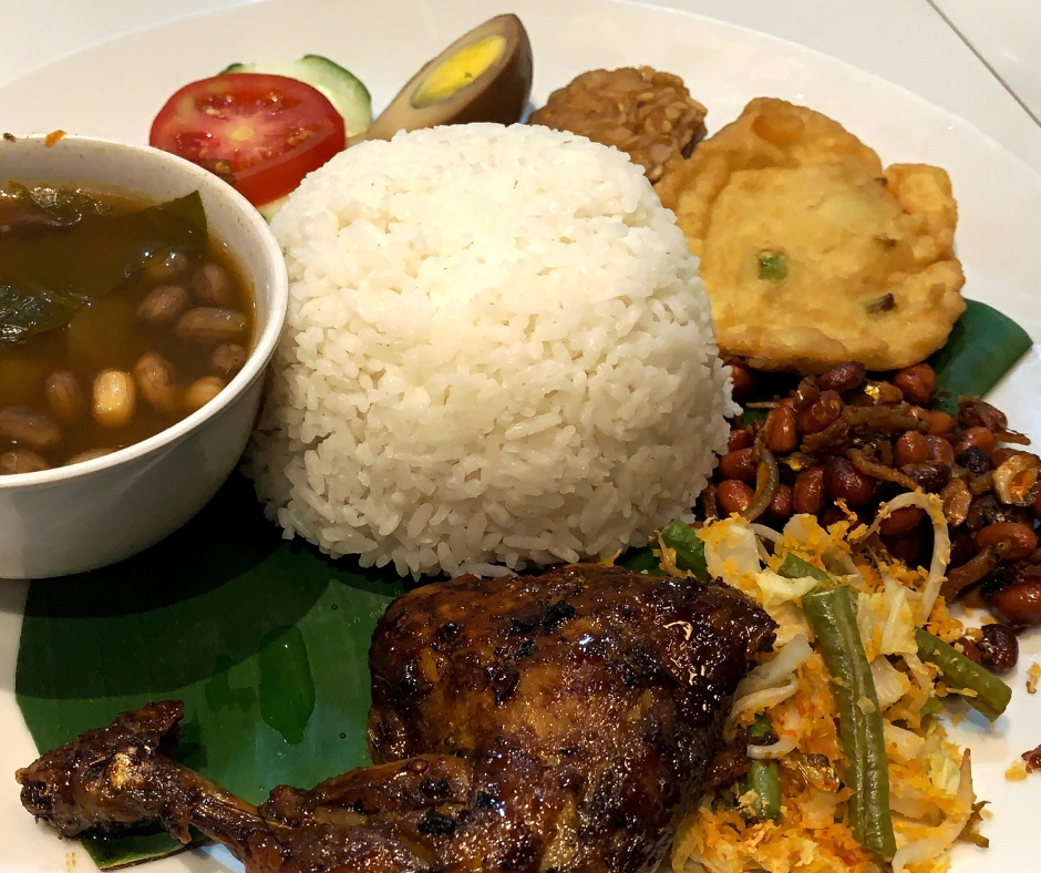 Traditional Bajan meal with grilled chicken, rice, peas, fried plantain, vegetables, and soup on a plate.