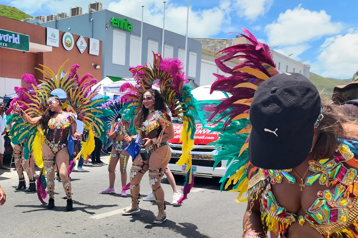 Masqueraders in pink, teal, and gold feathered costumes with jeweled bodysuits walking the parade route at SXM Carnival in St. Maarten
