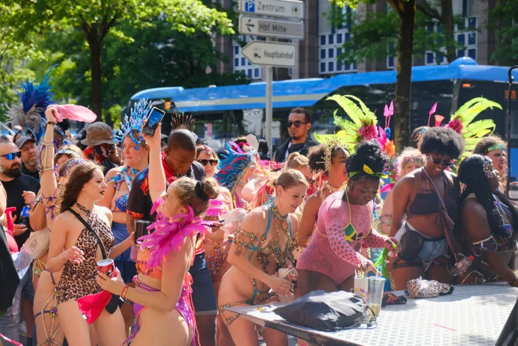 Masqueraders in feathered headpieces and gem costumes gathered at a drinks table during the Munich Carnival road march