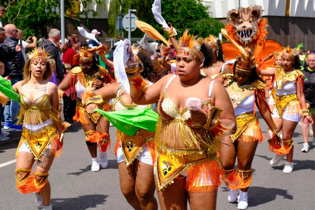 Masqueraders in gold and orange costumes with feathered headbands dancing through the streets during the Luton Caribbean Carnival procession