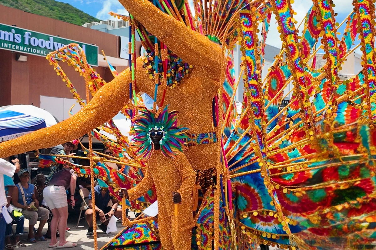 Masquerader in a towering gold glitter costume with rainbow tie-dye wings and gold sequin trim parading past spectators at St. Maarten Carnival.