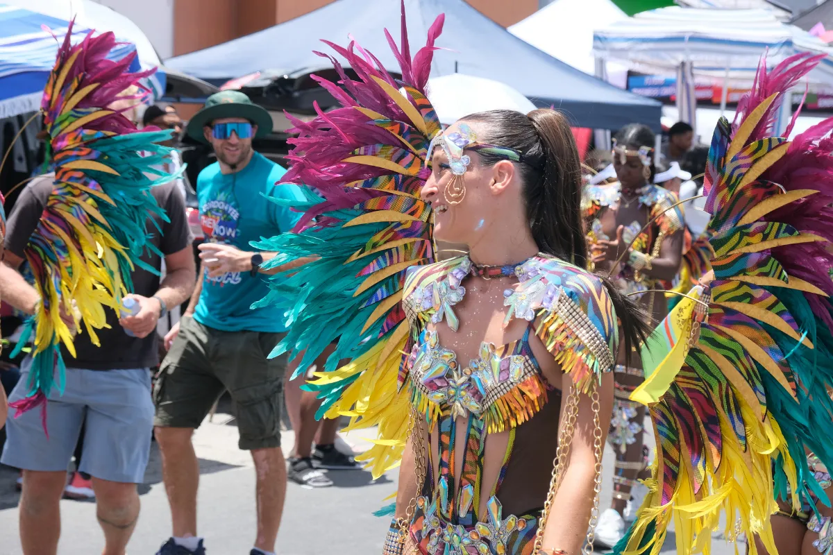 Smiling masquerader in a pink, teal and gold feathered costume with a spectator watching behind her