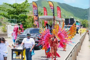 Masquerader in a red and pink feathered frontline costume walking the road at St. Maarten Carnival ahead of her band's music truck and drinks cart