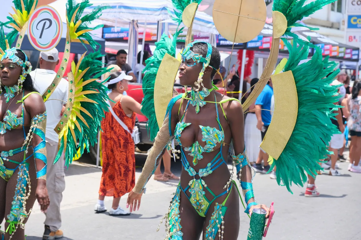Masquerader in a green and gold costume with feathered wings and a band cup in hand on the parade route at St. Maarten Carnival