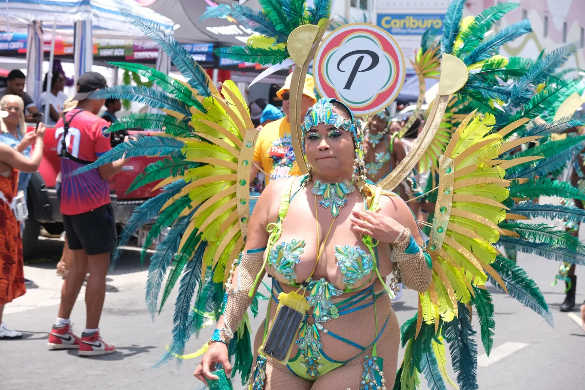 Masquerader in a green and gold feathered costume walking behind her section sign at Caribbean Carnival