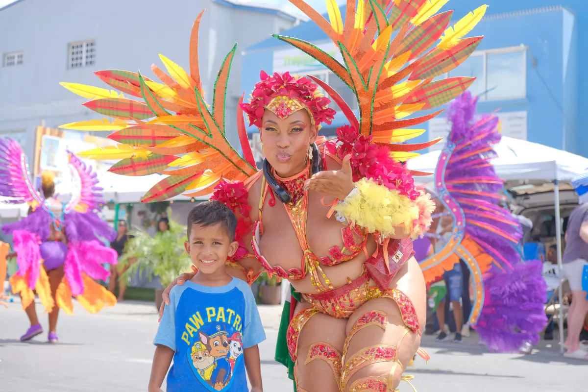 SXM Carnival masquerader, Lovely in a red, orange and yellow costume posing with a child along the parade route