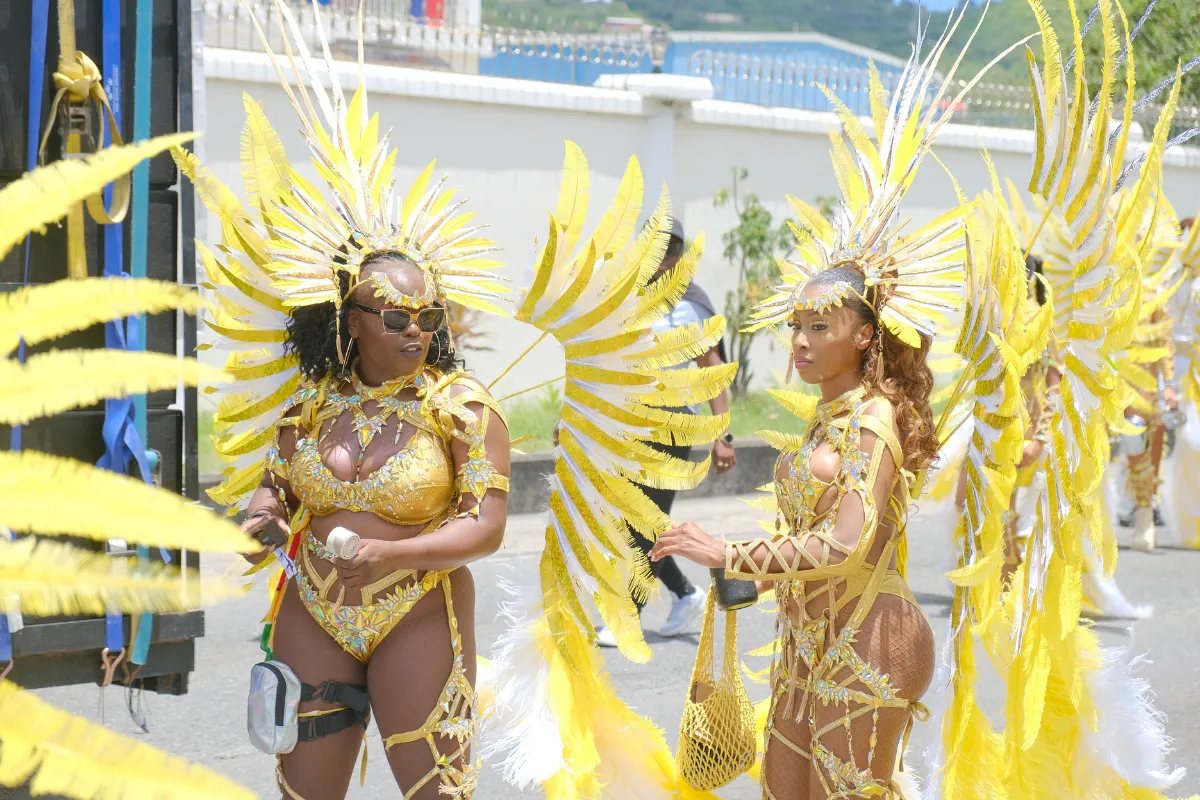 Two masqueraders in yellow and gold feathered costumes standing beside a music truck on Carnival day