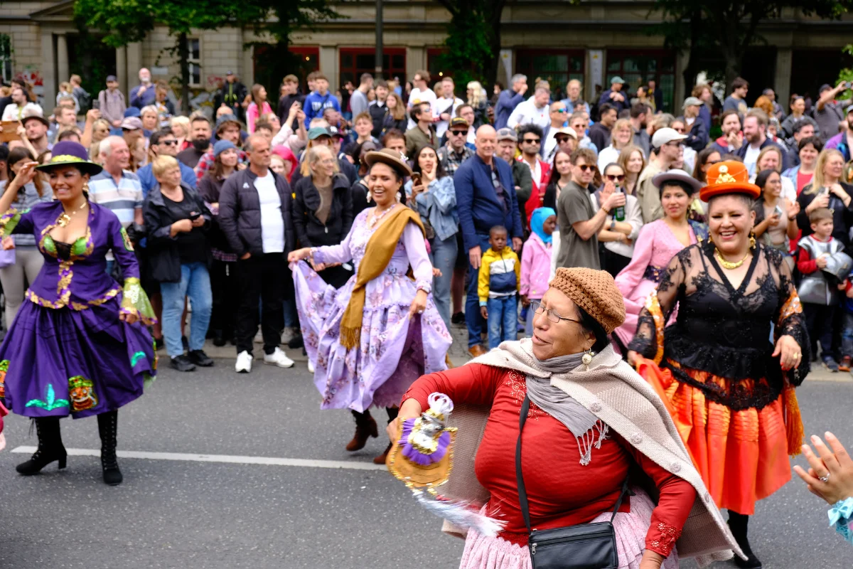 Carnival performers in purple lavender and orange traditional dresses dance through a crowded Berlin street as spectators watch