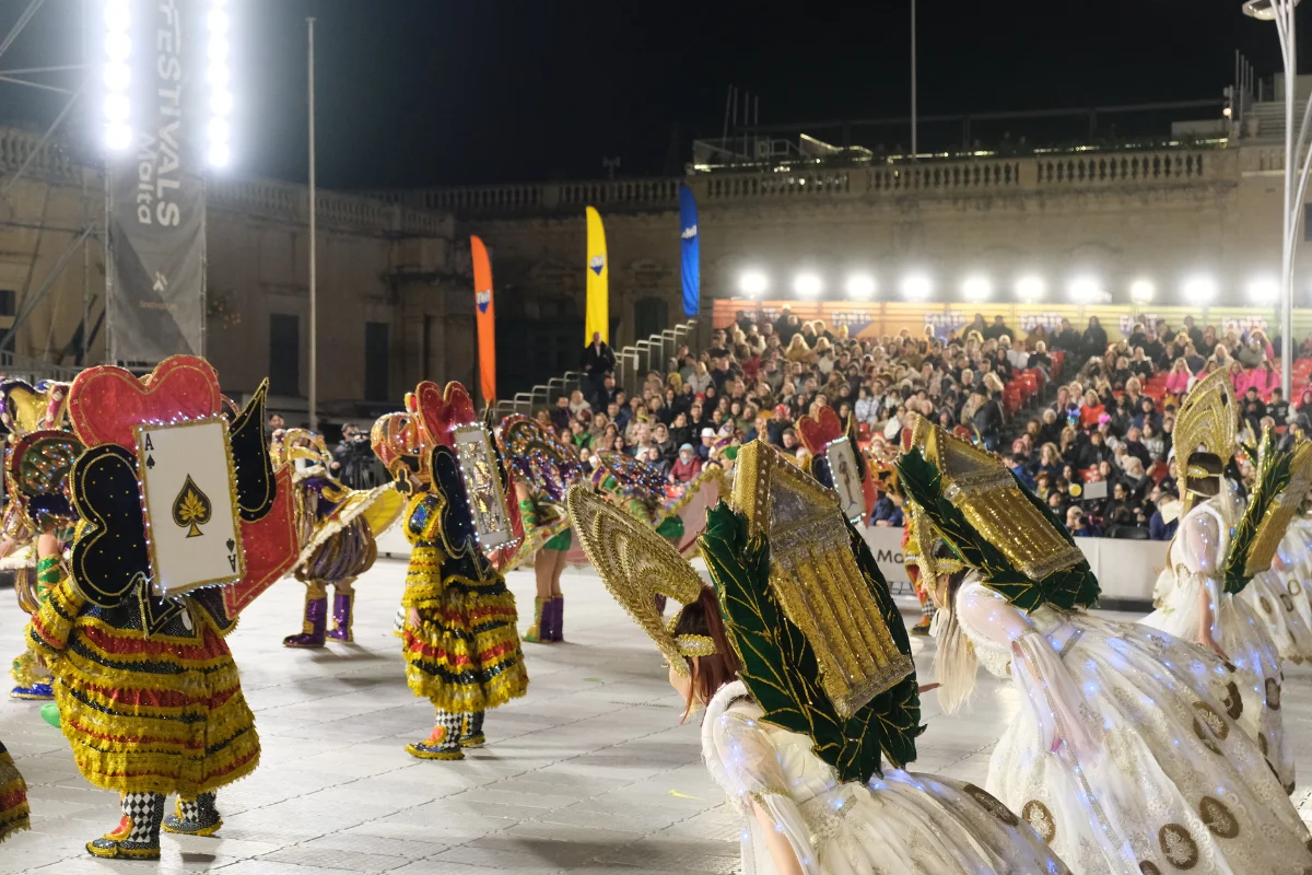 Dance company in card-themed costumes performing at an evening Malta Carnival show, with spectators seated in the stands