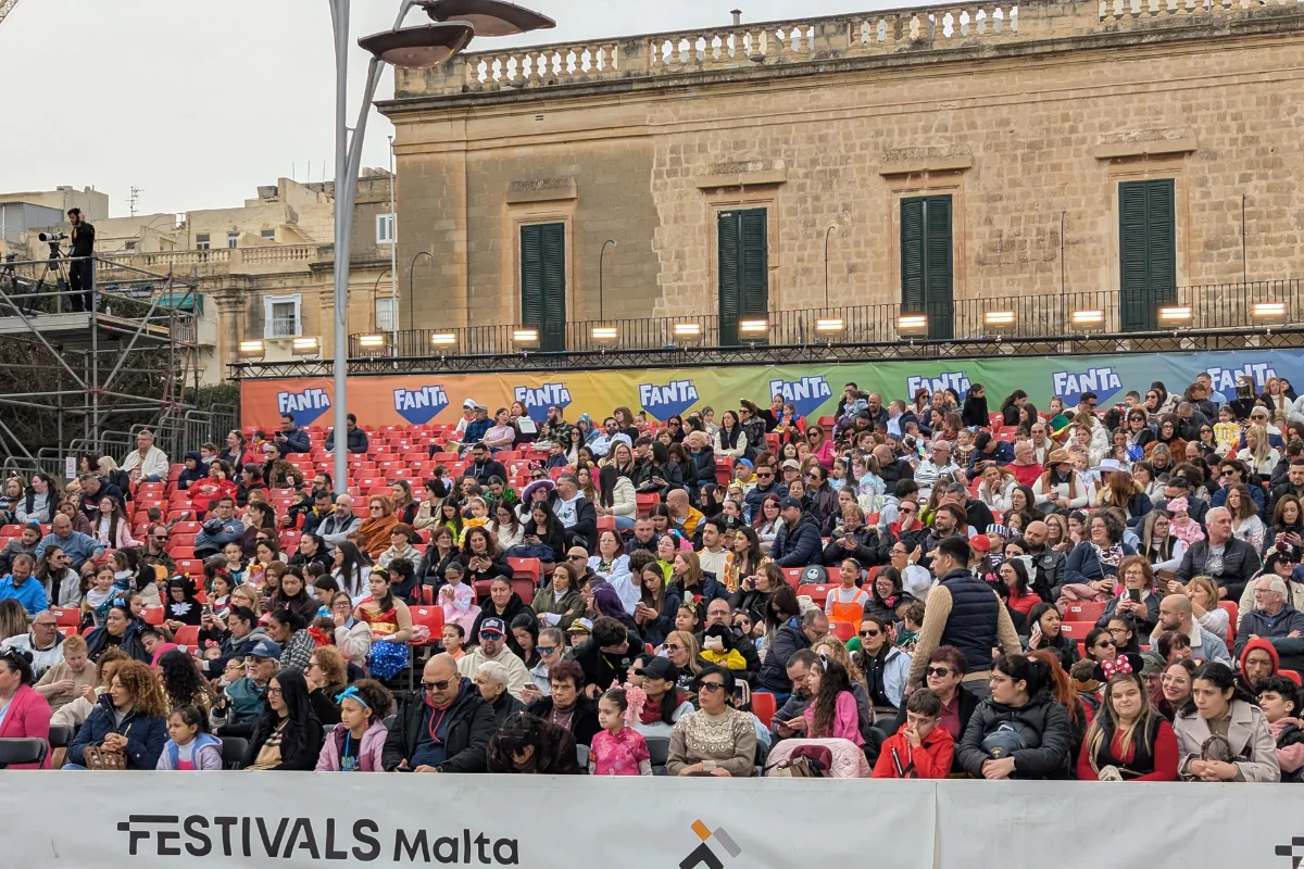 Families seated in the stands at a daytime Malta Carnival show, with children in costumes and adults in jackets