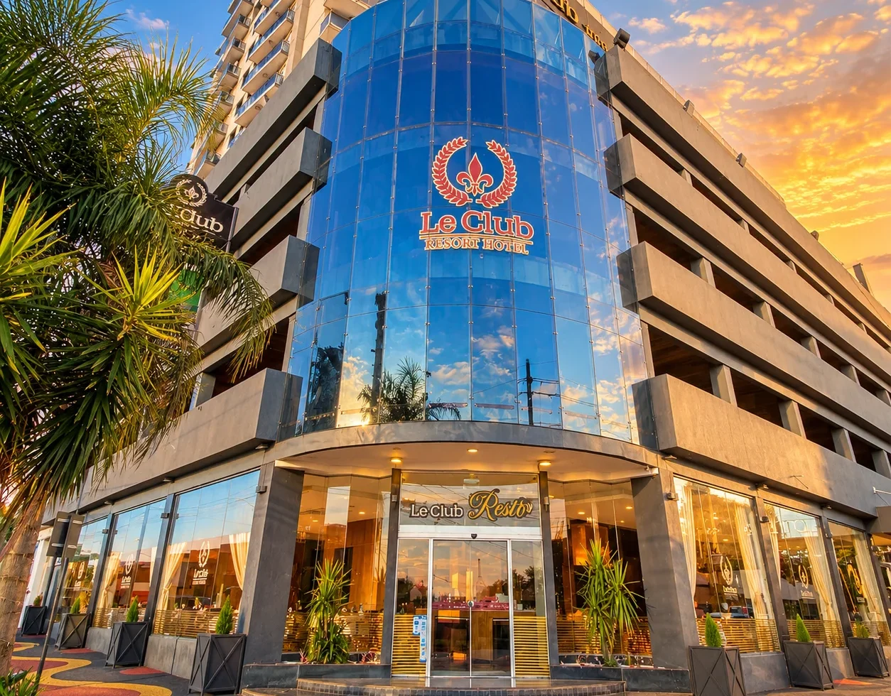 Le Club Resort Hotel exterior with a curved blue glass facade palm trees and illuminated entrance at sunset