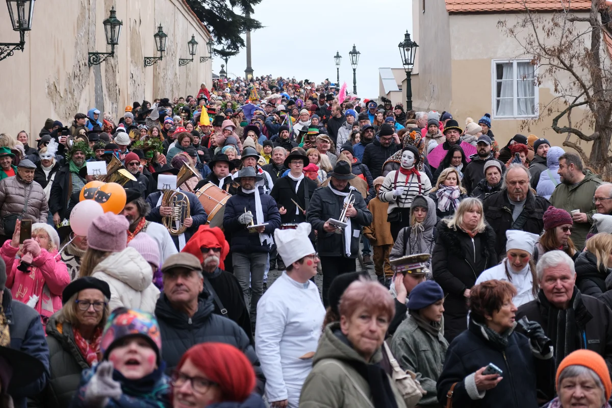 A large crowd of costumed revellers and spectators fills a narrow historic street during Prague Carnival with musicians playing brass instruments