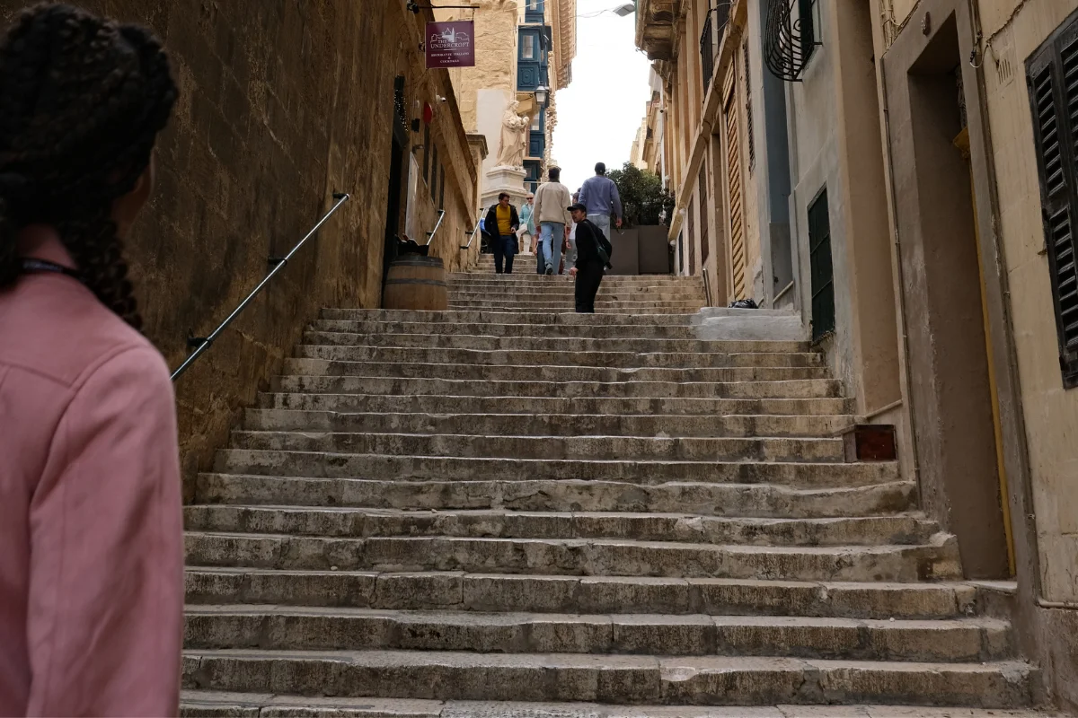 Carnival Vibez blogger in a pink jacket at the foot of a long stone staircase street in Valletta