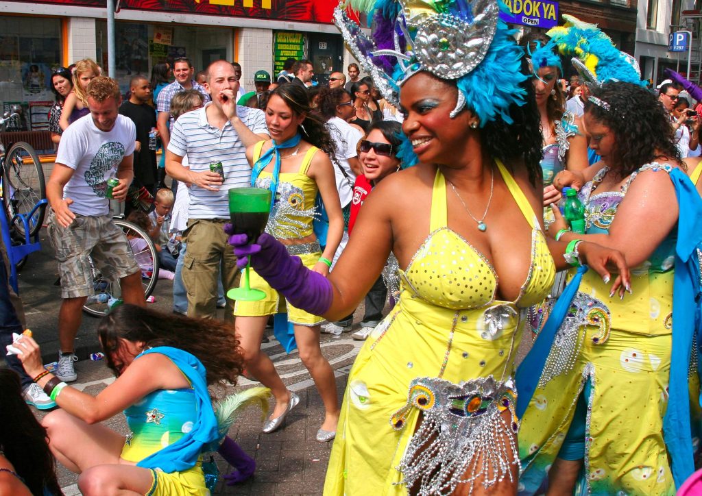 Woman in Carnival costume dancing down the streets at Rotterdam Zomercarnaval with a happy crowd in the background dancing along on the sidewalks.